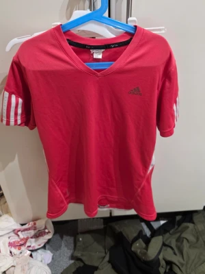Röd Adidas t-shirt med vita ränder - Snygg röd t-shirt från Adidas med klassiska vita ränder på ärmarna och svart Adidas-logga på bröstet. Tröjan har v-ringad hals och är gjord i ett lätt och ventilerande syntetmaterial, perfekt för träning eller sport.