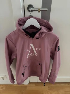 Rosa skidjacka från Slade Couture - Säljer en snygg rosa skidjacka från Slade Couture med huva och stor ficka framtill. Jackan har tryckta loggor både fram och bak samt på ärmen. Insidan är fodrad med mjuk fleece för extra komfort. Perfekt för dig som vill ha en stilren och funktionell jacka.