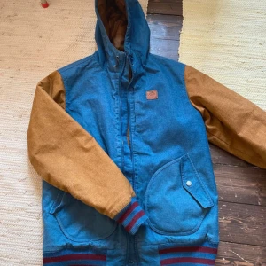 Blå och bruna vindjacka med huva - Snygg vindjacka i blått med bruna ärmar och huva. Jackan har dragkedja framtill, två stora fickor och ribbade muddar med rödblåa ränder. Perfekt för dig som gillar streetstyle och vill ha en jacka som sticker ut.