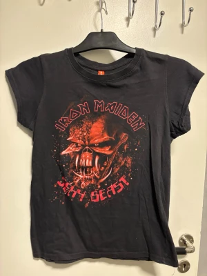 Iron Maiden svart t-shirt - Säljer en svart Iron Maiden t-shirt med ett fett tryck av ett monster och texten 'Iron Maiden' och 'Sexy Beast' i rött. T-shirten har korta ärmar och är gjord i mjuk bomull. Perfekt för dig som gillar rock och vill sticka ut.