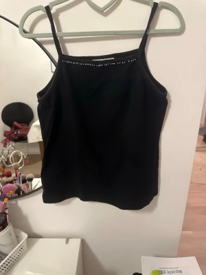 Svart linnetopp med pärlor från Friday - Svart linnetopp från Friday med smala axelband och dekorativa pärlor längs halsringningen. Toppen är enkel och stilren, gjord i mjukt bomullsmaterial. Perfekt för dig som gillar minimalistisk design med en liten twist.