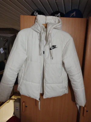Vit pufferjacka vändbar från Nike stl S - Säljer en riktigt snygg vit pufferjacka från Nike med stor svart logga på ryggen och mindre logga på bröstet. Jackan har huva med snörning och dragkedja framtill. Perfekt för kalla dagar och ger en clean look. Jackan är vändbar 
