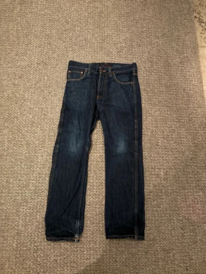 Mörkblå jeans från Nudie Jeans - Säljer ett par mörkblå jeans från Nudie Jeans i klassisk rak modell. Selvedge jeans som kostar 1800kr nya men som säljs här för ett rabatterat pris. Modellen är slim Jim. Jeansen har orangea sömmar, fem fickor och snygga detaljer på bakfickorna. Tillverkade i jeansmaterial och passar perfekt till en avslappnad stil.