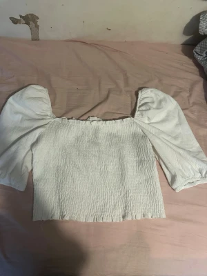 Vit blus med puffärmar - Säljer en söt vit blus med puffiga långärmar och rynkad resår vid axlarna. Toppen har en croppad passform och är gjord i ett lätt, strukturerat tyg som känns luftigt och skönt. Perfekt för dig som gillar romantisk och trendig stil.