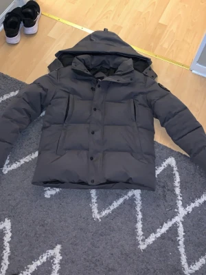 Grå pufferjacka från Canada Goose - Säljer en grå pufferjacka från Canada Goose med huva och flera fickor framtill. Jackan har en tydlig logotyp på ärmen och är fodrad för extra värme. Perfekt för kalla dagar och har en stilren look som funkar till det mesta.