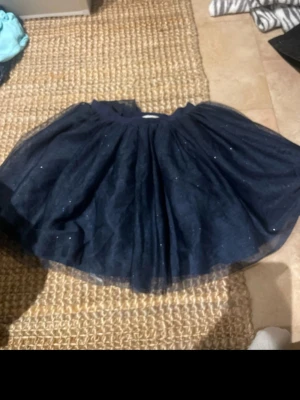 Mörkblå glittrig tyllkjol barn  - Säljer en fluffig mörkblå kortkjol i tyll med glittriga detaljer. Kjolen har en bred resår i midjan och flera lager som ger extra volym. Perfekt för dig som vill sticka ut med lite extra glitter och volym i outfiten.