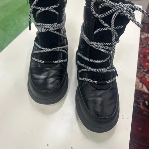 Svarta vinterstövlar från Kamik - Svarta vinterstövlar från Kamik med tjockt foder och snörning i svartvitt mönster. Yttermaterialet är syntet och skinn, med quiltade detaljer och mjuk insida. Perfekta för kalla dagar och har en platt sula för stabilitet.