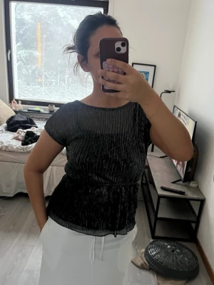 Glittrig svart topp från Vero Moda - Säljer en svart glittrig topp från Vero Moda i storlek M. Toppen har korta ärmar, rund halsringning och ett lätt figurnära snitt. Materialet är glansigt med silvertrådar som ger en snygg shimmer-effekt. Perfekt för dig som vill sticka ut lite extra.