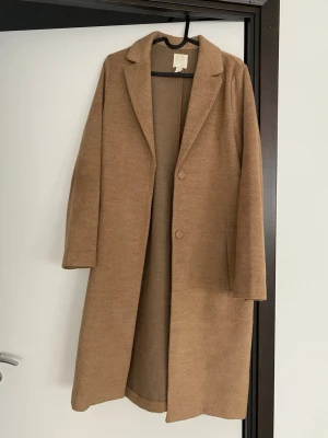 Beige kappa med klassisk krage - Snygg beige kappa från h&m med klassisk krage och två knappar framtill. Långärmad och med fickor på sidorna. Perfekt för dig som gillar stilrena och tidlösa plagg. Materialet känns mjukt och har en ullig look.