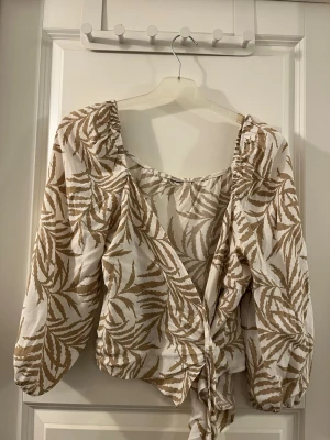 Beige och vit omlottblus med knyt - Säljer en snygg omlottblus i beige och vitt med ett zebrainspirerat mönster. Blusen har långa ärmar med lätt puff och knyt framtill. Materialet känns lätt och luftigt, perfekt för varmare dagar. Passformen är normal och blusen har en v-ringad hals.