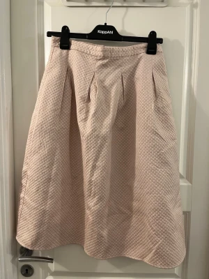 Ljusrosa långkjol från H&M, strl 38 - Säljer en ljusrosa långkjol från H&M i storlek 38. Kjolen har ett diskret vävt mönster med lite glansiga trådar som ger en snygg textur. Modellen är A-linjeformad med veck upptill för extra volym och fall. Perfekt för dig som gillar mjuka färger och stilren design.