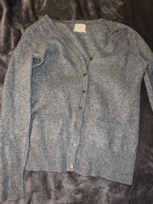 Grå kofta i 100% kashmir  - Mysig grå kofta i kashmir! Koftan har v-ringning, knappar hela vägen, två fickor framtill och långa ärmar. Perfekt för lager på lager och riktigt skön mot huden. Enkel och stilren design som funkar till allt. 100% Kashmir 