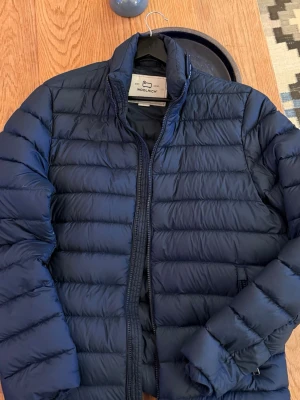 Woolrich dunjacka  - Säljer en stilren och lätt dunjacka från Woolrich. Relativt tunn men varm.  Marinblå färg. Klassisk passform. Fylld med dun (85% dun, 15% fjäder).  Sparsamt använd, inga hål eller fläckar. Dragkedja och sömmar i gott skick.