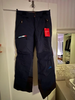 Mörkblå täckbyxor från Elevenate - Säljer ett par mörkblå täckbyxor från State of Elevenate i storlek medium. Byxorna har flera dragkedjeförsedda fickor, förstärkta partier vid bensluten och coola röda detaljer på dragkedjorna. Perfekta för vinterns alla äventyr. Nya med tags kvar.