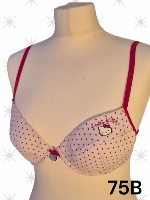Hello Kitty prickig bh-topp 75B - Supersöt bh-topp från Hello Kitty i vitt med röda prickar och tunna röda axelband. På ena kupan finns ett Hello Kitty-tryck och en liten rosett med Hello Kitty-hänge framtill. Perfekt för dig som gillar gulliga detaljer och lekfull stil. 