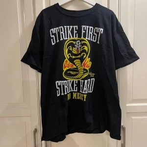 Svart T-shirt med tryck - Svart t-shirt med Cobra Kai-tryck på framsidan och texten 'Strike First, Strike Hard, No Mercy'. I 100% bomull. Endast testad.