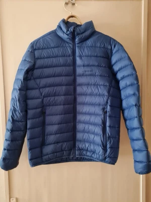 Blå dunjacka från Everest (Stadium) strl S - Blå dunjacka från Everest (Stadium) i storlek S. Jackan har horisontella quiltade sektioner, hög krage och två sidofickor med dragkedja och har innerficka. 
