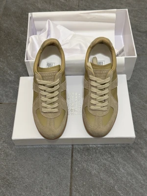 Maison Margiela beige sneakers - Maison Margiela sneakers i beige med klassisk låg siluett. Skorna har ovandel i mocka och skinn, beige snörning och gummisula i brunt. Tidlös design med rund tå och diskreta detaljer, perfekt för dig som gillar minimalistisk stil.