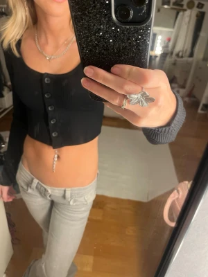 Svart topp  - Säljer en svart croppad topp från Gina Tricot i ribbad bomullsmix. Toppen har långa ärmar, knappar framtill och en figurnära passform. Perfekt till jeans eller kjol för en trendig look.