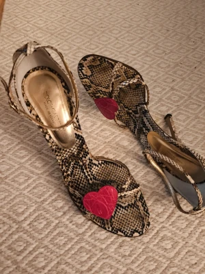 Dolce & Gabbana sandaler med hjärtdetalj - Unika sandaler från Dolce & Gabbana i äkta ormskinnsmönstrat skinn med smala remmar och hög klack. Framtill sitter en röd hjärtdetalj som ger extra edge. Färgerna är beige, svart och röd. Tillverkade i Italien med exklusivt material och snygg form. Nypris 7000-8000