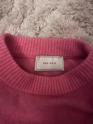 Rosa stickad tröja från Neo Noir - Mysig rosa stickad tröja från Neo Noir med rund hals och ribbade muddar. Tröjan har puffade axlar och långa ärmar, vilket ger en cool och trendig siluett. Perfekt för att addera färg till din garderob under kyliga dagar.