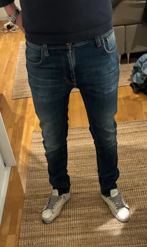Nudie Jeans - Säljer ett par Nudie jeans i mörkblå tvätt med snygga slitningar. Jeansen har orange sömmar och en smal passform som sitter tajt men ändå bekvämt. Jeansen har tecken på användning men är annars i fint skick! Tveka inte att höra av dig om du har några frågor eller prisförslag!