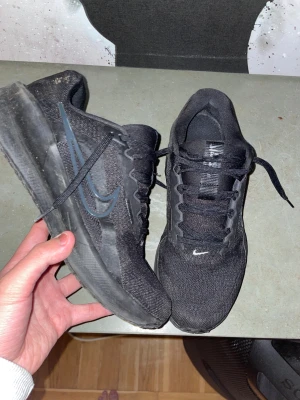 Svarta Nike skor - Säljer ett par svarta Nike löparskor med klassisk swoosh på sidan. Skorna är i väldigt bra skick, en snabbt tvätt får dem att se ut som nya. platt sula och snörning. Perfekta för träning eller till vardags. Skorna är väldigt bekväma.