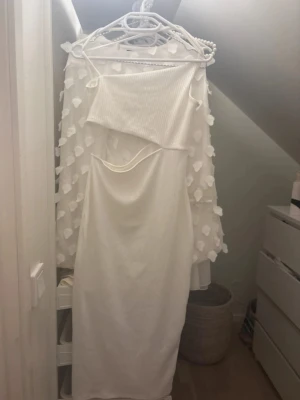 Vit ribbad långklänning - Säljer en vit långklänning från H&M i ribbad polyester.  Klänningen har en öppning på sidan. Klänningen har en axelbandslös stil och ett coolt snitt vid midjan som ger en modern vibe. Perfekt för dig som gillar trendiga och stilrena plagg.