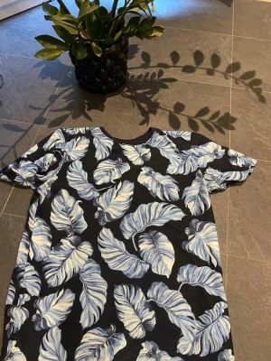 Svart t-shirt med blått bladmönster Lindbergh - Snygg svart t-shirt från Lindbergh med stort blått och vitt bladmönster över hela plagget. T-shirten har rund hals och korta ärmar, perfekt för dig som gillar tropiska vibes och vill sticka ut lite extra. Materialet känns mjukt och skönt mot huden.