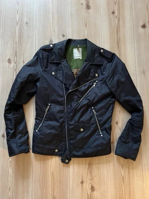 Svart bikerjacka från J.Lindeberg - Svart bikerjacka från J.Lindeberg i storlek S, tillverkad i 100% polyester. Jackan har flera dragkedjor, snedställd frontzip, axelklaffar och coola detaljer i silver. Perfekt för dig som gillar en edgy stil och vill sticka ut lite extra.