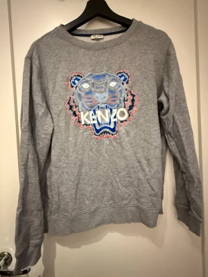 Grå Kenzo sweatshirt  - Säljer en grå sweatshirt från Kenzo med ikonisk broderad tiger i blått, vitt och rött framtill. Tröjan har rund hals, långa ärmar och är gjord i mjuk bomull. Perfekt för dig som vill ha en statement piece i garderoben. Storlek 16 Kids ca storlek S/M 