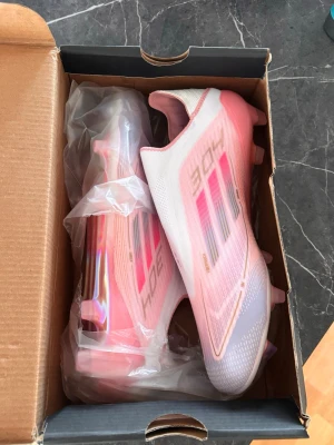 Adidas F50 rosa fotbollsskor - Säljer ett par Adidas F50 fotbollsskor i vitt och rosa med metallicdetaljer. Skorna har en strömlinjeformad design utan snörning, tre klassiska ränder på sidan och en cool lila innersula. Ovandelen är i syntetmaterial och sulan har fasta dobbar för gräs.