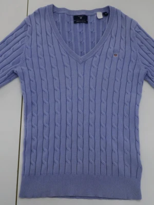 Ljuslila kabelstickad tröja från Gant - Snygg ljuslila kabelstickad tröja från Gant med v-ringad hals och korta ärmar. Tröjan har ett litet broderat Gant-emblem på bröstet och är gjord i mjuk bomull. Perfekt för dig som gillar klassisk stil med en modern twist.