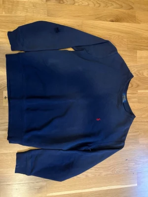 Mörkblå sweatshirt från Polo Ralph Lauren - Säljer en mörkblå sweatshirt från Polo Ralph Lauren med klassisk röd broderad logga på bröstet. Tröjan har rund hals, långa ärmar och är gjord i mjuk bomull. Perfekt för dig som gillar stilrena och tidlösa plagg det enda är att de är ett litet hål i armen därför säljer jag den billigt!