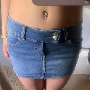 Blå jeanskjol med fickor - Säljer en blå jeanskjol i klassisk denim med två framfickor och två bakfickor. Kjolen har dubbla silverknappar i midjan och bälteshällor. Snygg kort modell som är enkel att matcha med olika toppar. Perfekt för dig som gillar en chill och trendig stil.