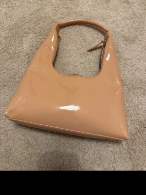 Beige axelväska i blank syntet - Snygg beige axelväska med stilren, fyrkantig form och blank finish. Väskan har ett rejält handtag och stängs med dragkedja. Insidan har ett litet fack för småsaker. Perfekt för dig som gillar minimalistisk design och vill ha en väska som sticker ut.