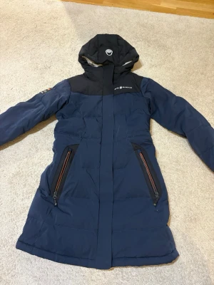 Marinblå dunjacka från Sail Racing - Säljer en marinblå lång dunjacka från Sail Racing som passar en XS. 