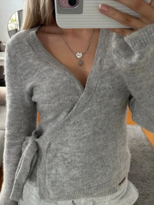 Grå omlottkofta från Gina Tricot - Supermysig grå omlottkofta från Gina Tricot i ullblandning. Koftan har långa ärmar, v-ringning och knytband i sidan som ger en snygg passform. Perfekt att slänga på sig när det är lite kyligt ute. Ribbstickade muddar vid ärmslut och nederkant.