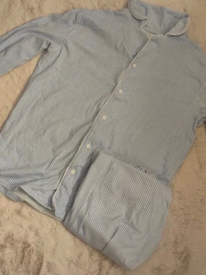 Blåvit randig pyjamas med knappar! - Klassisk pyjamas i två delar med blå och vita tunna ränder. Skjortan har långa ärmar, vit knappstängning och en mjuk krage. Byxorna har resår i midjan för extra komfort. Perfekt för mysiga kvällar hemma!