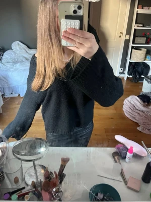 Svart stickad tröja med v-ringning - Säljer en svart stickad tröja med bred v-ringning och långa, vida ärmar. Tröjan har en avslappnad passform och ribbade muddar vid ärmslut och nederkant. Perfekt för lager på lager och enkel att styla till olika outfits. ytterst lite nopprig