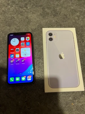 Lila iPhone 11 med originalkartong - Säljer en snygg lila iPhone 11 från Apple. Telefonen har en stilren design med rundade kanter, glasbaksida och dubbla kameror. Kommer med originalkartong. Perfekt för dig som vill ha en modern och färgglad mobil.