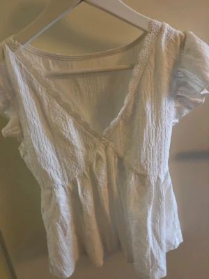 Vit spetsblus med volangärmar - Söt vit blus med v-ringning och spetsdetaljer. Blusen har korta volangärmar och ett broderat mönster i tyget. Perfekt för dig som gillar romantisk och feminin stil. Lätt och luftig, passar till många olika outfits.