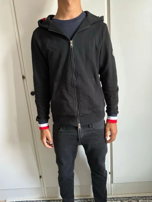 Svart Moncler hoodie med dragkedja - Säljer en svart hoodie från Moncler med hel dragkedja framtill och huva. Tröjan har Moncler-logga på vänster ärm och ribbade muddar med röd, vit och blå rand. Klassisk och stilren design, perfekt till streetwear-looken. Materialet känns mjukt och bekvämt.