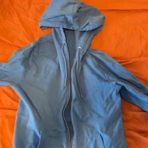 Blå hoodie med dragkedja - Säljer en blå hoodie med dragkedja framtill och justerbar huva med snören. Hoodien har en avslappnad passform och är tillverkad i mjukt bomullsliknande material. Perfekt för en chill och bekväm stil.