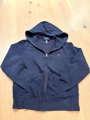 Mörkblå hoodie från Polo Ralph Lauren - Mörkblå hoodie med dragkedja från Polo Ralph Lauren. Klassisk design med huva, kängurufickor och röd broderad logga på bröstet. Tillverkad i mjukt bomullsmaterial som är skönt att ha på sig. Perfekt för en avslappnad och stilren look.  Hör av dig vid minsta fundering😊