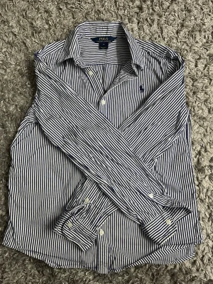 Randig skjorta från Polo Ralph Lauren - Snygg skjorta från Polo Ralph Lauren i vit och blå med smala vertikala ränder. Klassisk krage, långärmad och med knappar framtill. Broderad logga på bröstet. Tillverkad i mjuk bomull som känns skön mot huden.