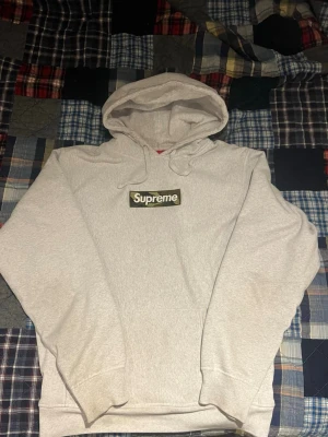 Supreme grå hoodie med camo box logo - Supreme hoodie i ljusgrått med klassisk box logo på bröstet i camo-mönster och vit text. Hoodien har huva med dragsko och en stor magficka. Perfekt för dig som gillar streetwear och vill sticka ut med en ikonisk look.