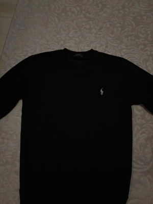 Svart tröja från Polo Ralph Lauren - Klassisk svart tröja från Polo Ralph Lauren med rund halsringning och den ikoniska broderade loggan i vitt på bröstet. Enkel och stilren design som passar till det mesta. Perfekt basplagg för garderoben.