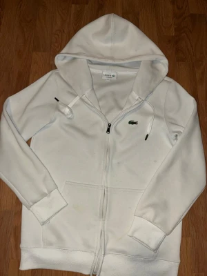 Vit zip-hoodie från Lacoste - Snygg vit hoodie från Lacoste med dragkedja och klassisk krokodil-logga på bröstet. Hoodien har huva med dragsko, ribbade muddar och fickor framtill. Perfekt för en clean och sportig look. Har några fläckar, Storlek S sitter perfekt. 
