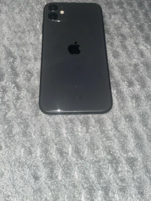 Svart iPhone 11 med dubbla kameror - Säljer en svart iPhone 11 med stilren glasbaksida och rundade kanter. Telefonen har två bakre kameror och Apples logga centrerad på baksidan. Skärmen är stor och täcker nästan hela framsidan, perfekt för sociala medier och streaming.